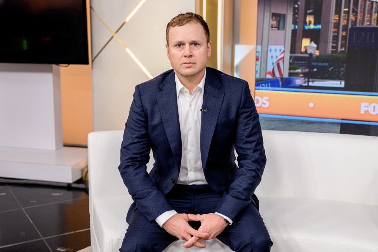 Spencer Pratt seated on a white couch with his hands folded in his lap.