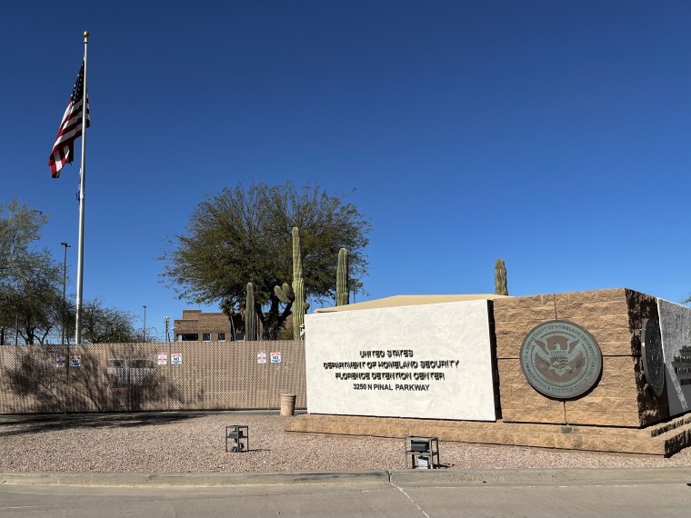 A stone sign outside a fenced area with cacti standing near an American flag on a pole.