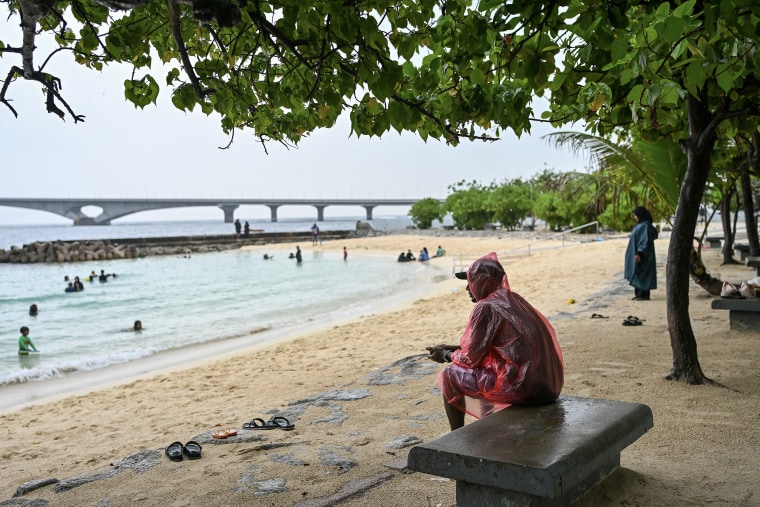 A man wearing a transparent red plastic parka sits on a concrete bench on a beach.