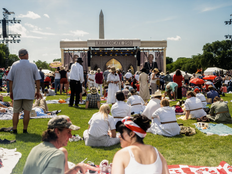 People sit on the grass and stand in front of an outdoor stage with the Washington Monument visible in the background.