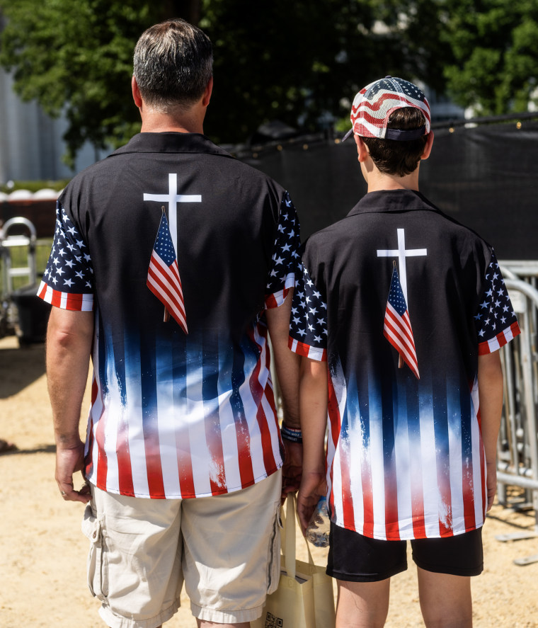 A father and son stand with their backs to the camera wearing polo shirts with a white cross overlayed by the American flag.