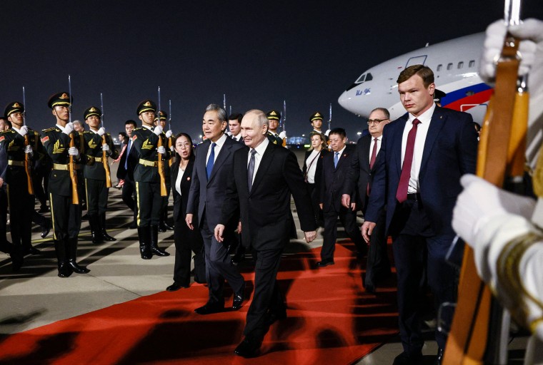 Vladimir Putin and Wang Yi walk along a red carpet extending from a plane parked in the background, with soldiers and other officials surrounding them