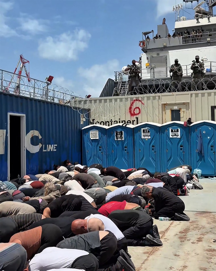 Detained activists kneeling in rows with their hands zip-tied behind their backs, in what appears to be an outdoor Israeli port facility.