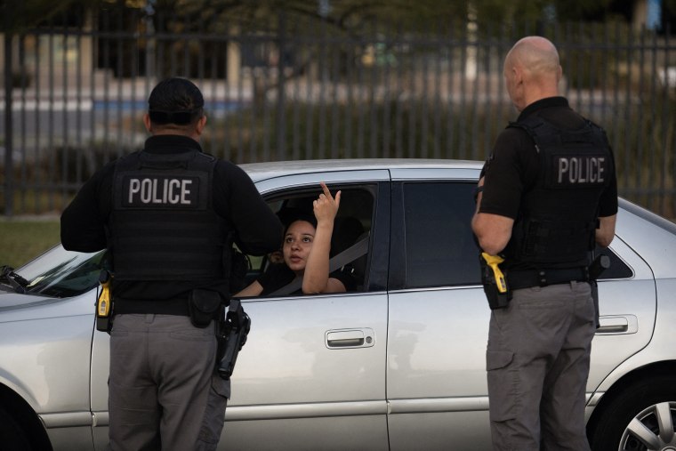 A woman seated in the driver's seat of a gray sedan speaks to two police officers standing outside the car.