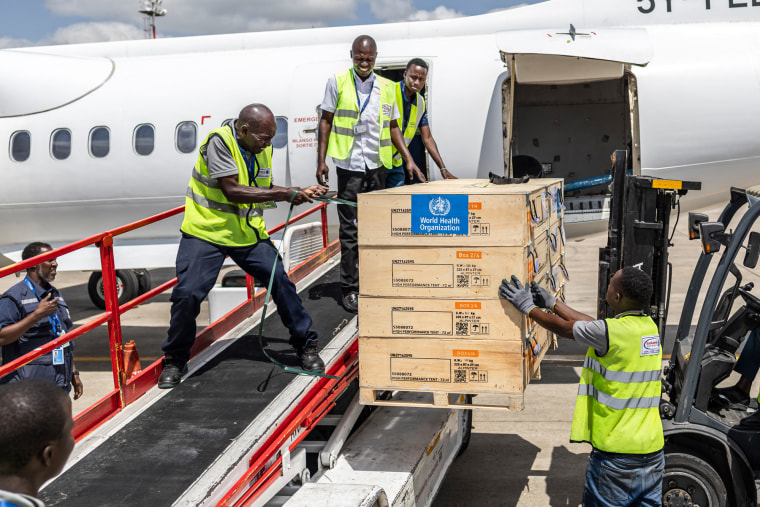 People wearing neon green vests load World Health Organization boxes onto a plane outside