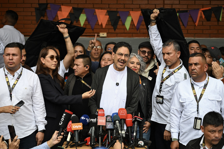 Ivan Cepeda standing at an array of microphones with aides and bodyguards surrounding him.