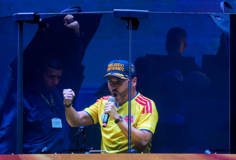 Abelardo de la Espriella speaks behind a wall of bulletproof glass, wearing a yellow soccer-style jersey and a blue baseball cap.