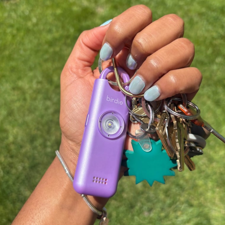 The Birdie safety alarm on a keychain while being held in a hand with blue fingernails against a grass background.