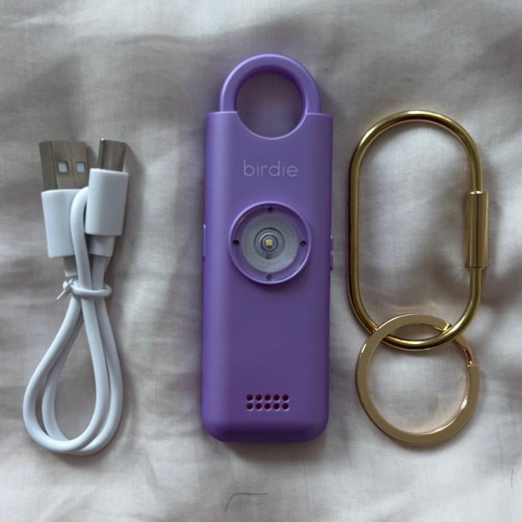 The Birdie USB-C charging cord, the Birdie alarm device and the gold keyring side by side on a pink bedspread.