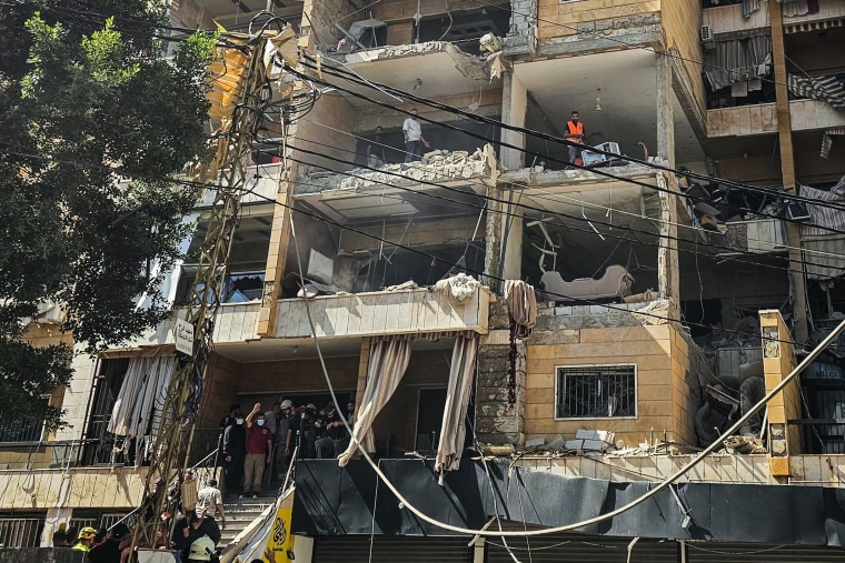People stand inside of the rooms of an apartment building with the exterior walls blown off