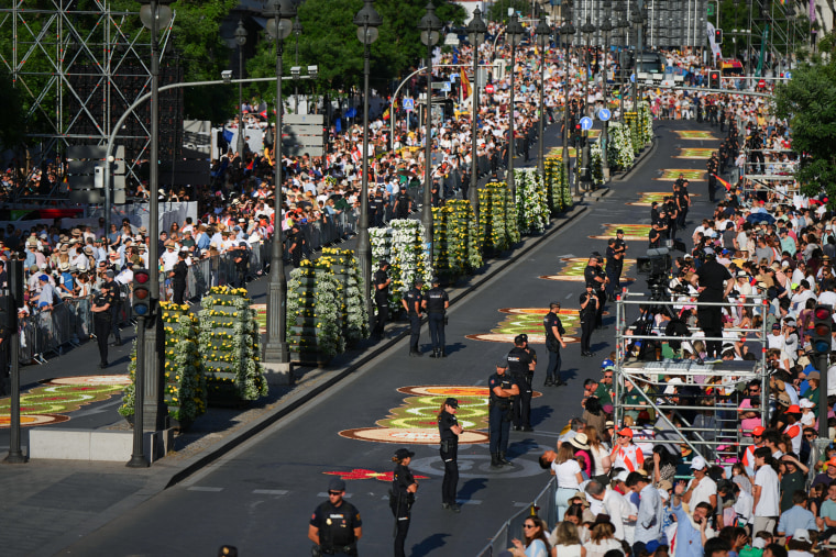 Flower petal beds can be seen on the street in between rows of people crowded on the sidewalks