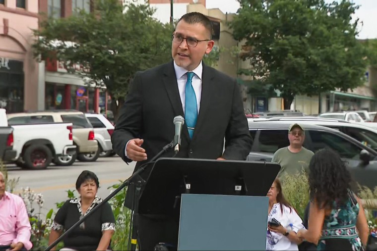 Roger Garcia speaks at a podium outside, facing away from the street, some people sit behind him