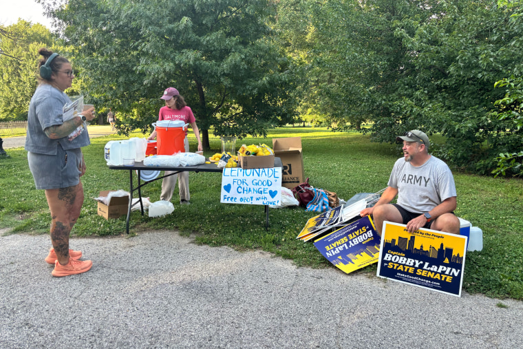 Bobby LaPin, right, seated on a cooler set on the grass, sits surrounded by Bobby LaPin campaign signs. Next to him is a lemonade stand, two women are also in the frame on the left side standing nearby