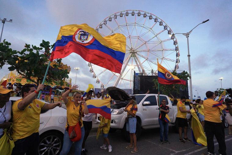 People wave Colombian national flags in front of a Ferris wheel.