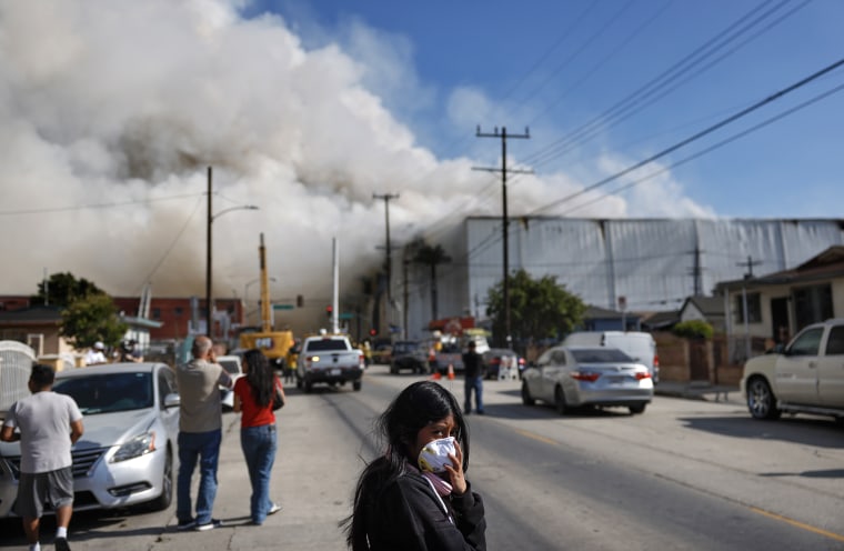 Thick smoke rises from an industrial building in Los Angeles as people gathered in the street look on.