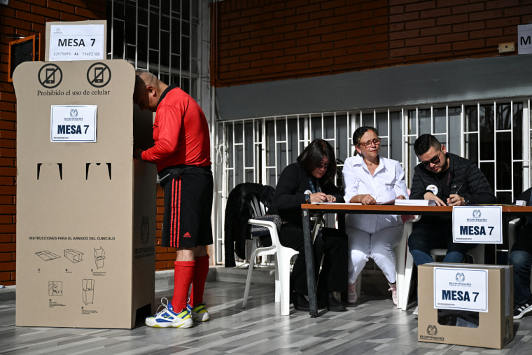 A man votes at a polling station booth next to a table of poll workers inside of a room