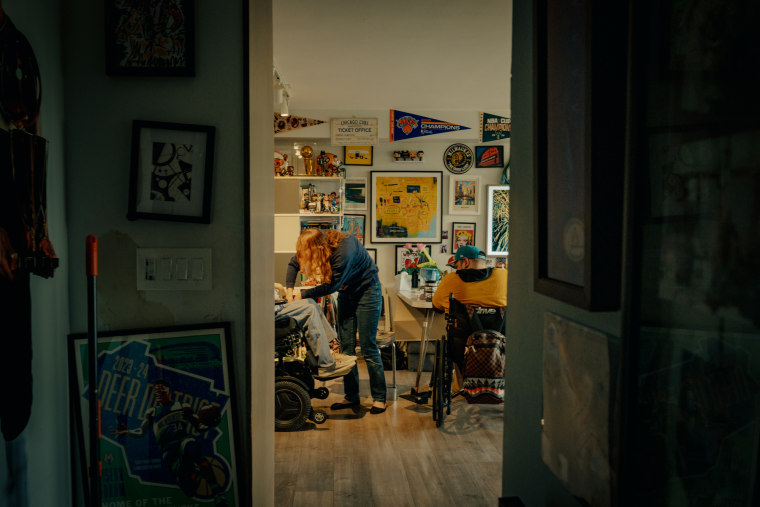 As seen through a doorway, a woman assists Rebecca Anger who is seated in a wheelchair, as Greg Anger watches, also seated in a wheelchair