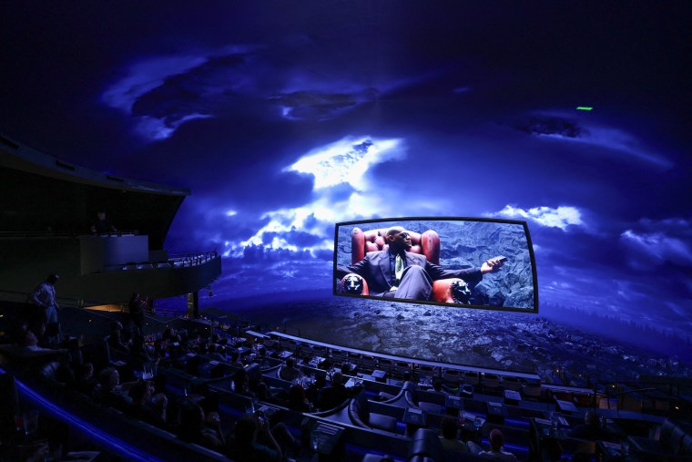 Attendees watch the character Morpheus appear on screen inside of a movie theater