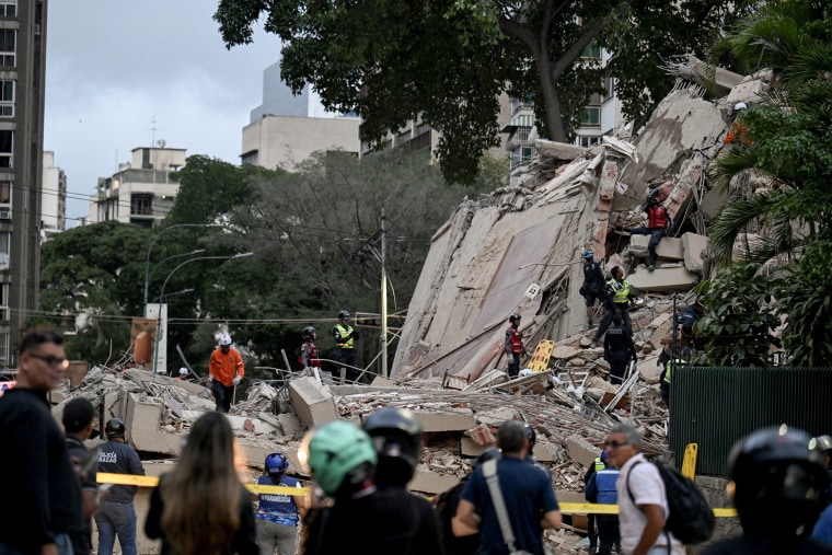 A crowd gathered behind yellow tape with a crumbled concrete building in the background.