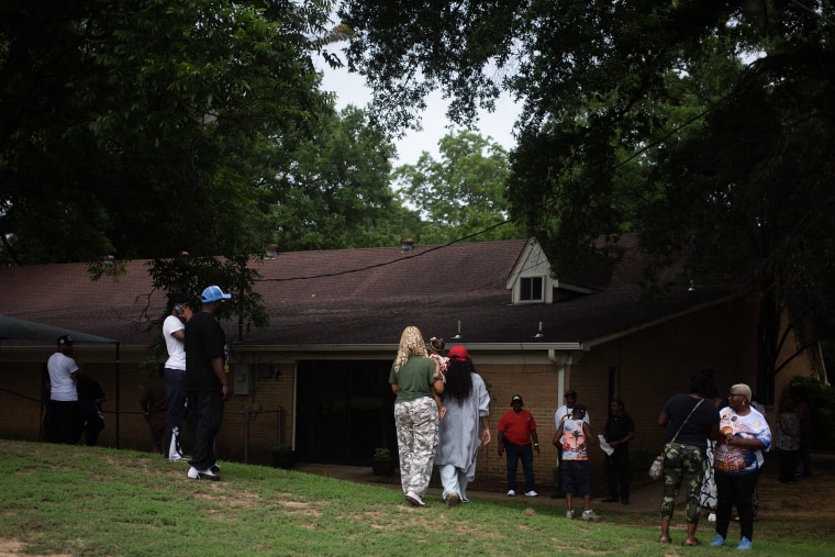 A group of people standing in the shade of tall trees in front of a brick building.