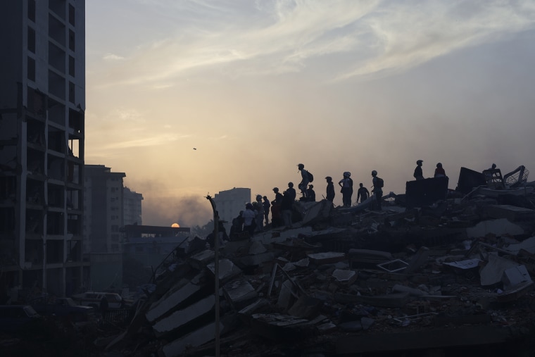 Rescue workers silhouetted by the setting sun stand atop a large pile of debris.
