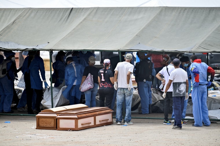 People gathered under a tent with white body bags lining the ground.
