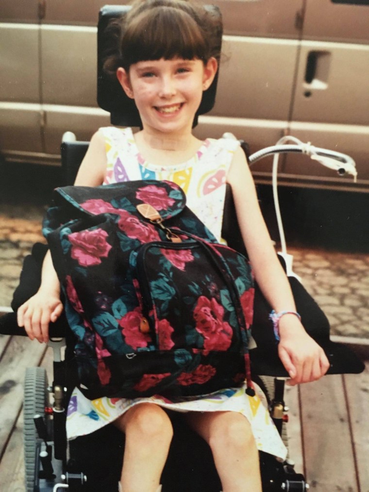 Rebecca Anger as a child, smiling, seated on a wheelchair with a floral design backpack on her lap