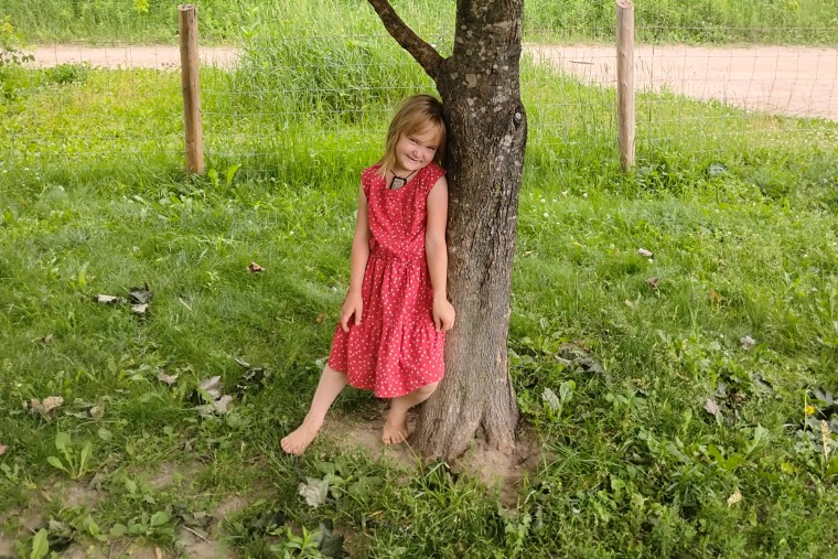 Cece Kale, a young girl, stands next to a tree while smiling to pose for a portrait, she is barefoot in the grass and wearing a pink dress with white polka dots