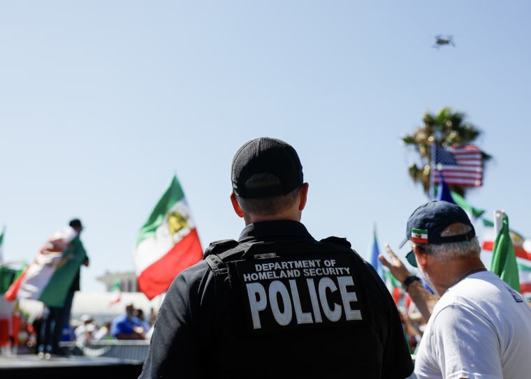 A DHS agent, seen from behind, looking up toward a drone in the sky.