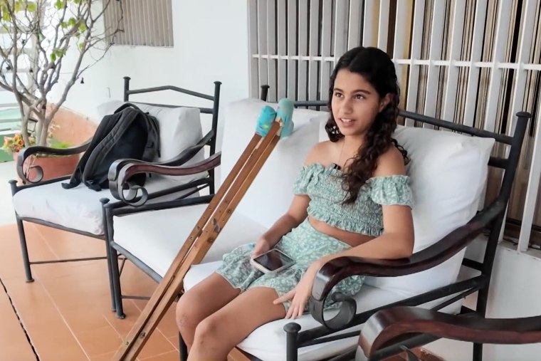 Fabiana Paso sits on a white-padded couch outside on a front porch, next to two crutches leaning up next to her