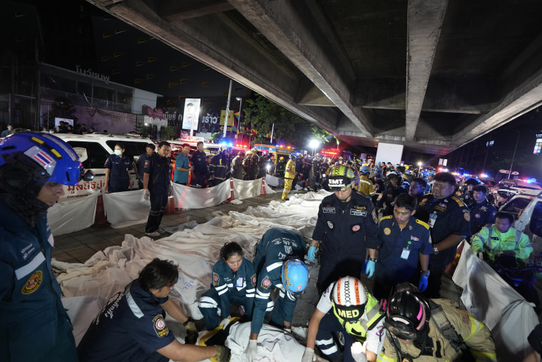 Emergency medical workers arrange a row of bodies covered in white sheets under a highway overpass at night.