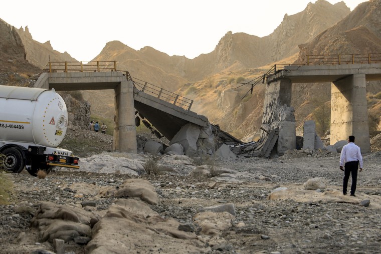A concrete bridge stands partially collapsed in an arid landscape as a man looks on.