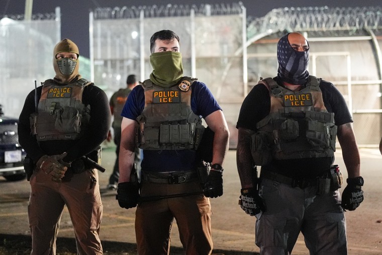 ICE officers wearing masks covering their noses and mouths stand outside near barbed wire fencing behind them