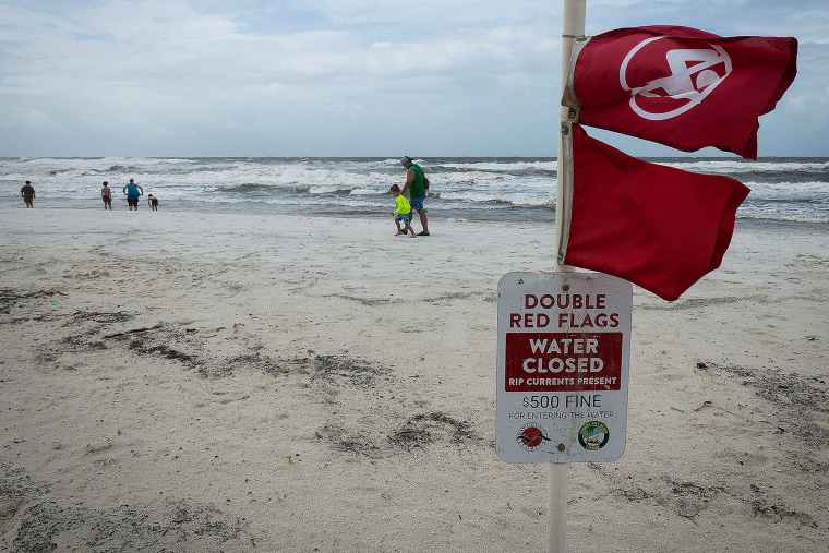 People walk by the shore on the beach, in the foreground are two red flags on a pole above a sign that reads "Double Red Flags, Water Closed, Rip Currents Present, $500 Fine for entering the water"