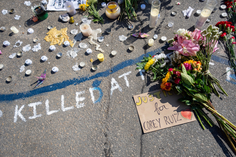 Velas, flores y un cartel de cartón que dice "Justicia para Corey Ruiz" se ven en una calle. Junto al cartel, escrito con tiza, se lee "Asesinado en...".