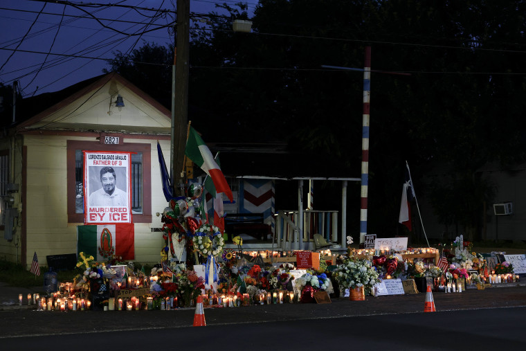 A makeshift memorial with dozens of candles and flowers laid in front of a small house at night.