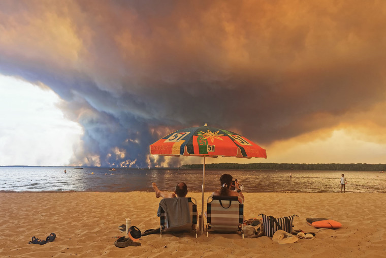 People relax on the beach in Lacanau, southwestern France, on July 24, 2026, as clouds of smoke rise into the sky from a wildfire. 