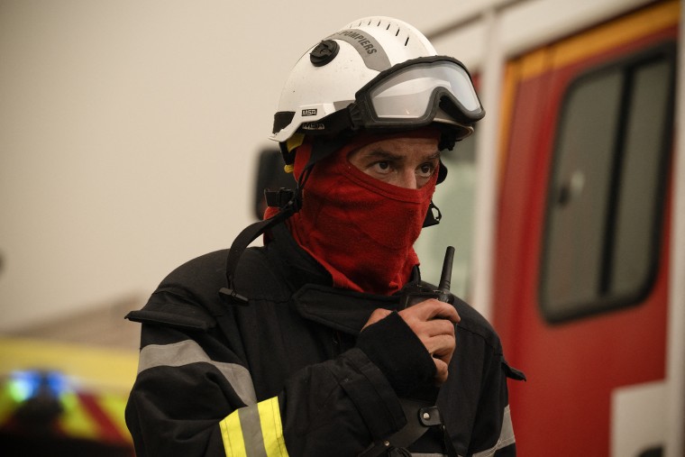 A close view of a firefighter wearing a hard hat and red face mask, holding a radio in his right hand.