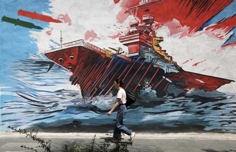 A young adult wearing earbuds walks in front of a war propaganda mural painted onto the side of a building.