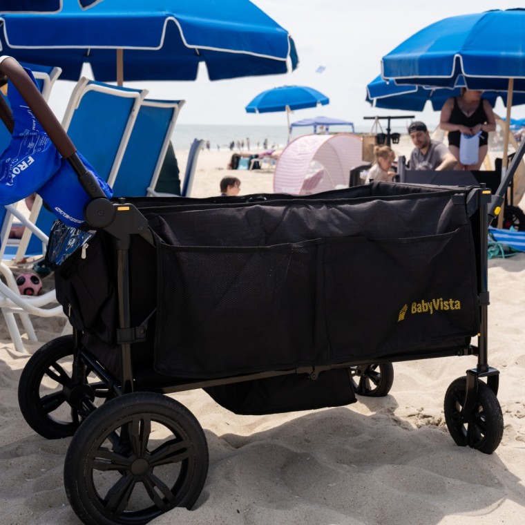 A black BabyVista folding wagon with mesh side pockets parked on sand at the beach, surrounded by blue umbrellas and beach chairs
