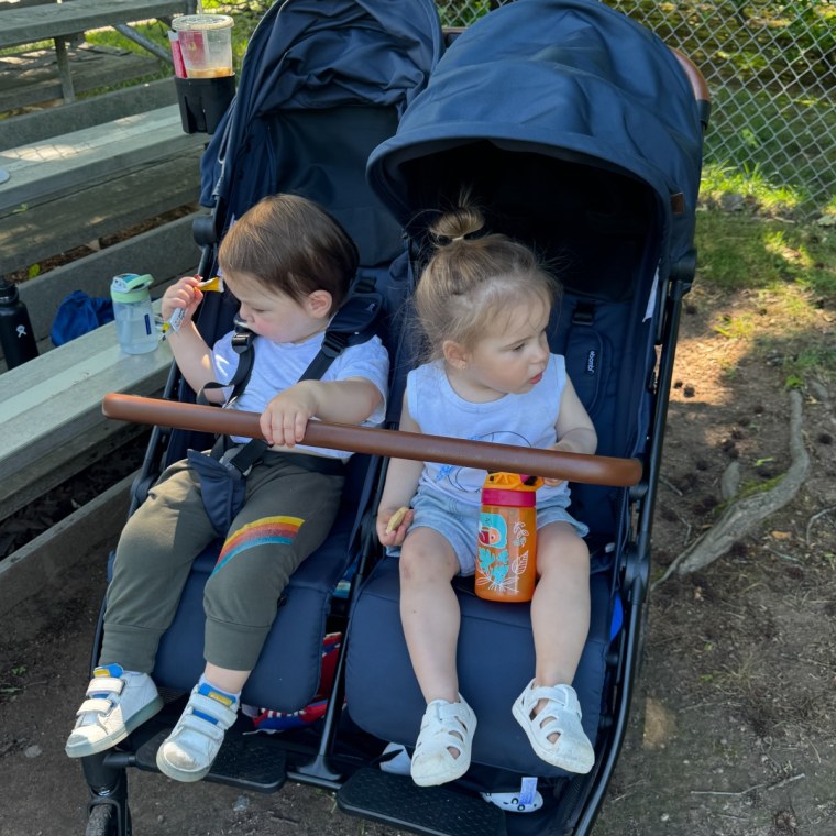 A toddler boy and girl sitting side by side in a navy blue Bombi double stroller with a wooden play bar, holding snacks and drinks outdoors near a chain-link fence