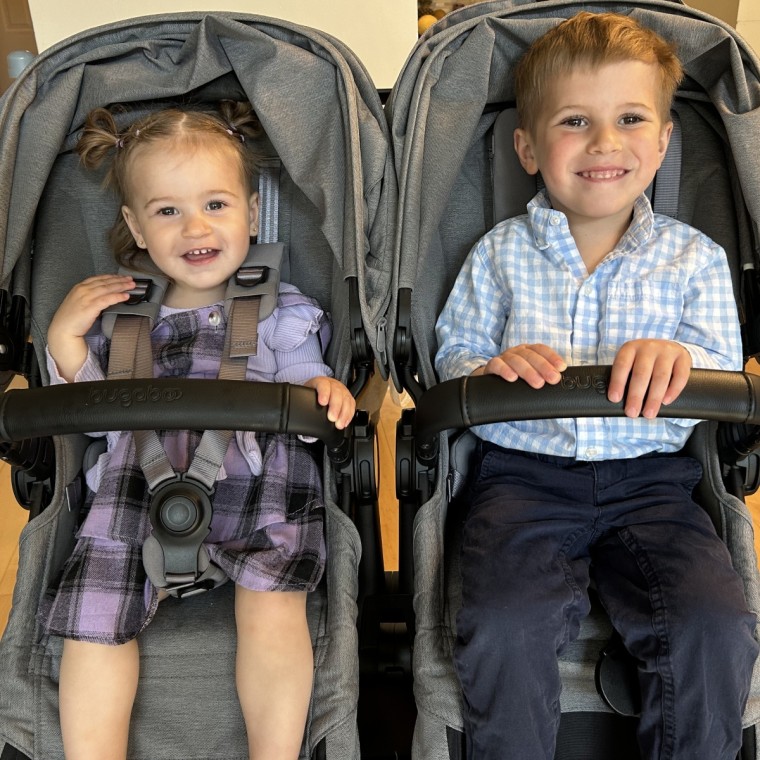 A smiling toddler girl in overalls and a young boy in a gingham shirt sitting side by side in a gray Bugaboo Donkey double stroller indoors, both holding the leather-trimmed handlebars