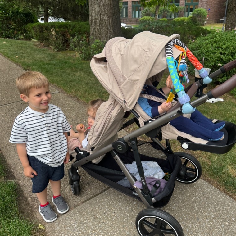 A smiling young boy standing next to a tan Cybex Gazelle S stroller with two toddlers seated inside, on a sidewalk lined with bushes and trees.