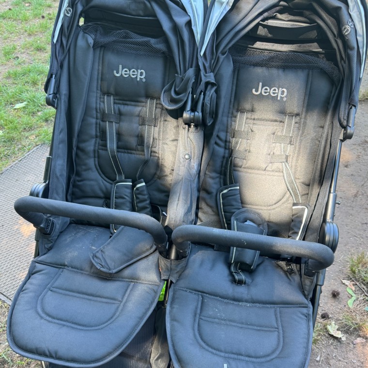 Overhead view of a black Jeep Destination Double stroller with two seats, harnesses, and canopies, sitting outdoors on a paved path next to grass