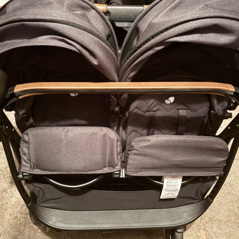 Close-up of a black Joie Rosemary LX double stroller's two canopies and seat backs, with a tan leather-trimmed handlebar and fabric storage basket below