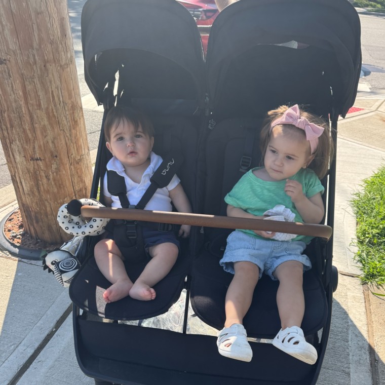 A baby boy and toddler girl with a pink bow sitting side by side in a black double stroller with a wooden play bar across the front, on a sidewalk