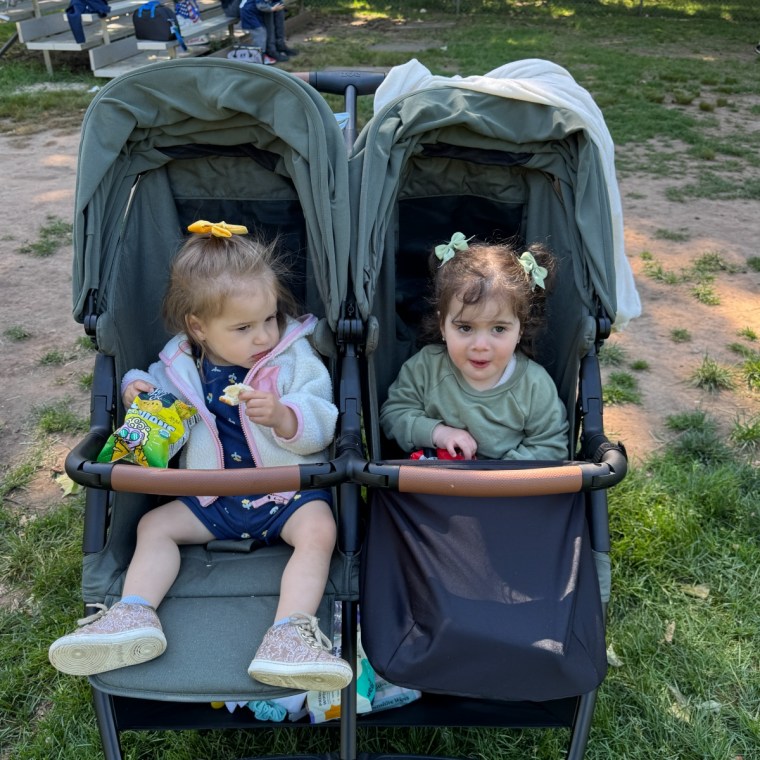 Two toddler girls with hair bows sitting side by side in a sage green double stroller at a park, one eating a snack from a bag
