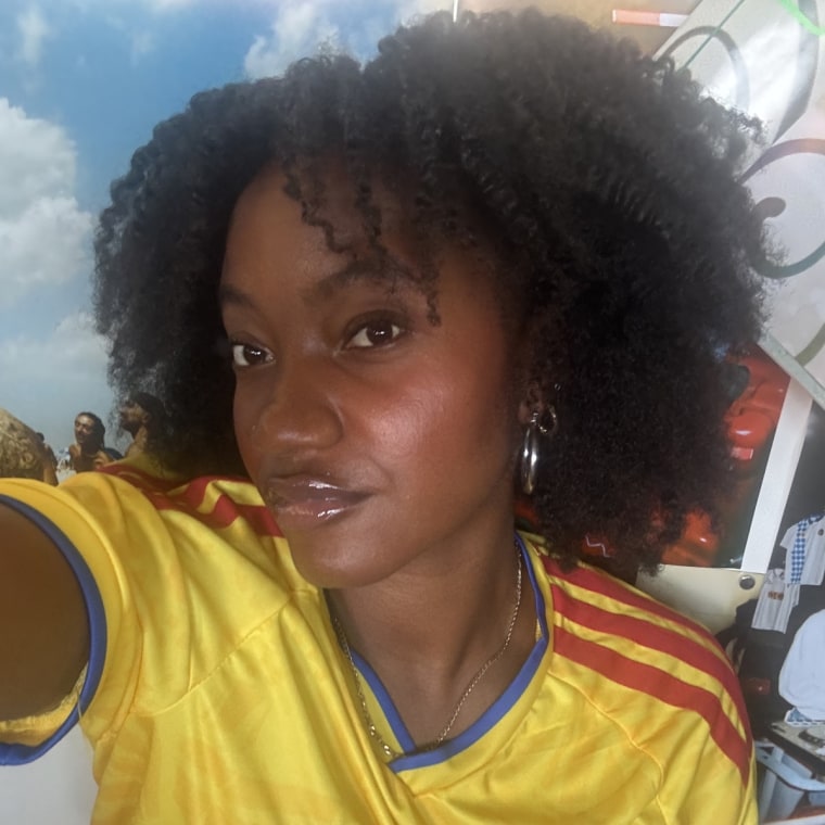 A selfie of Morris with a blown-out braidout using the Cecred Thermal Shield Mist, wearing a yellow soccer jersey in front of a wall of graffiti art.