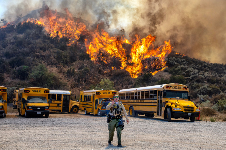 A brush fire is seen in the trees behind a row of parked school buses in a parking lot as a cop walks in the foreground speaking