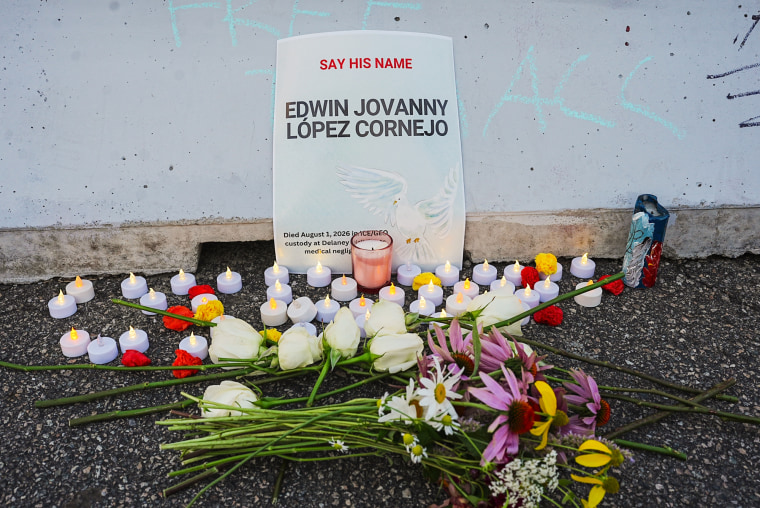Flowers and fake candles are placed on the ground in front of a placard leaned up against a wall that reads "Say His Name Edwin Jovanny Lopez Cornejo"